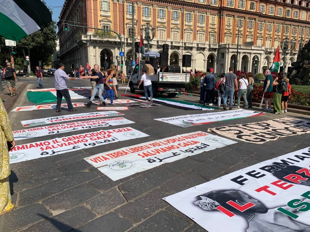 Photo of several banners and signs laid out on the ground in front of Piazza Statuto, bearing a variety of slogans in support of Palestine.