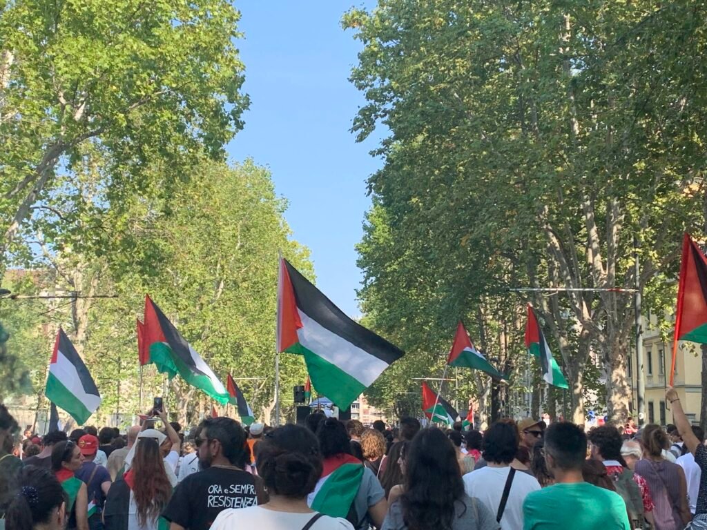 Photo taken from within the protest march, with trees on either side of the column. Several Palestinian flags are visible above the heads of the participants.