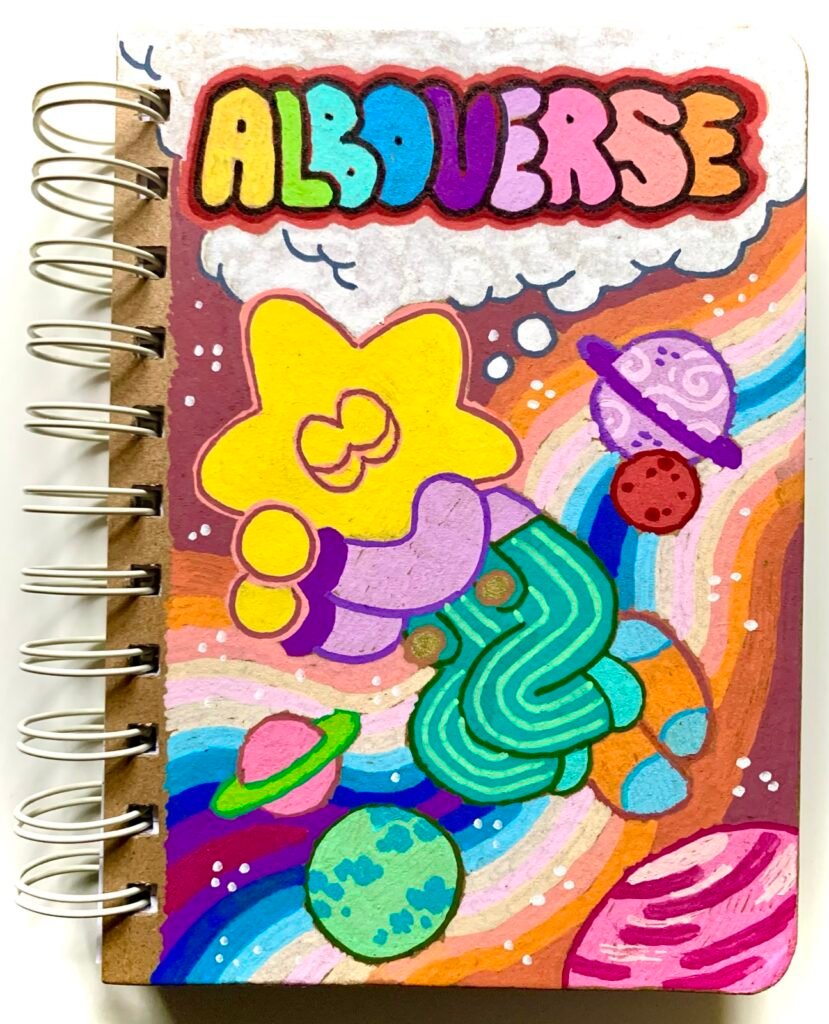 Photo of a notebook cover, decorated using bright posca pens. Below a rainbow coloured "Alboverse" header, is a sleeping star-headed toddler, surrounded by planets and stars.