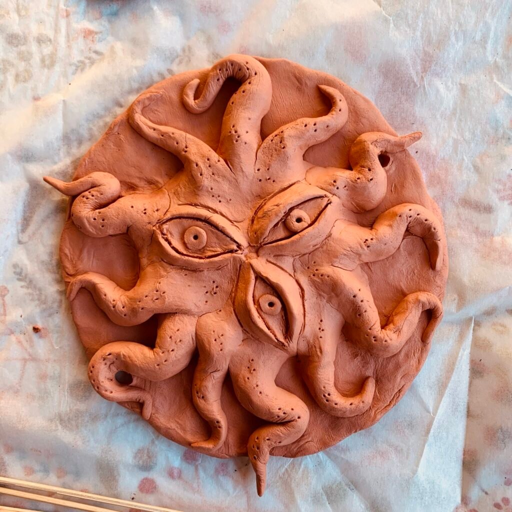 Photo of a hand-sculpted clay tablet, depicting a three-eyed tentacled creature, somewhat resembling a sun.